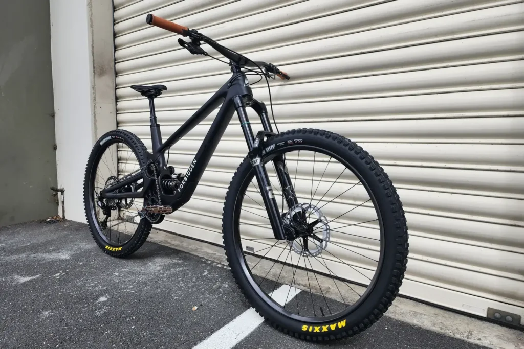 Black full-suspension mountain bike with tan grips, built by Outset, in front of a roller door
