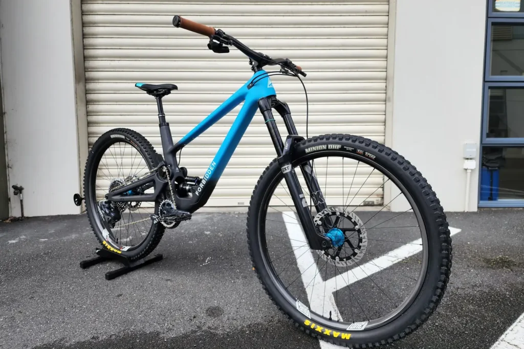 Blue full-suspension mountain bike built by Outset, on a stand outside the workshop